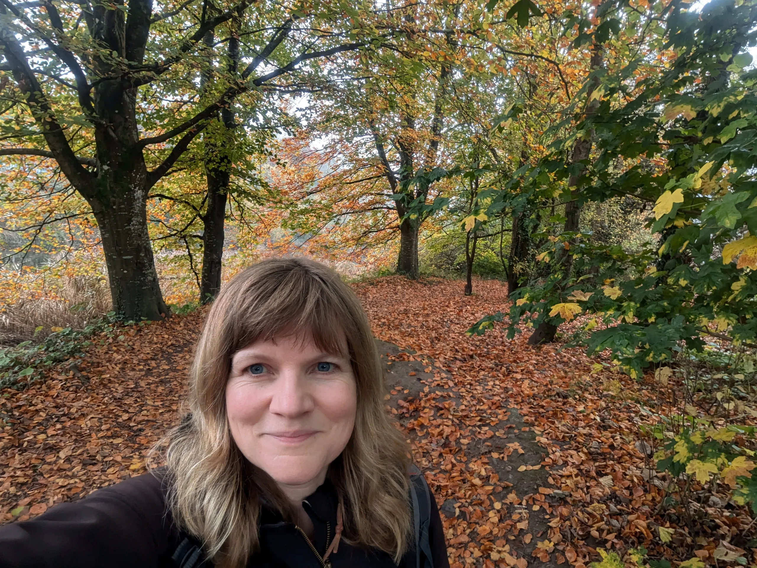 Smiling white woman in a black jumper standing in a woodland clearing with beech leaves on the ground