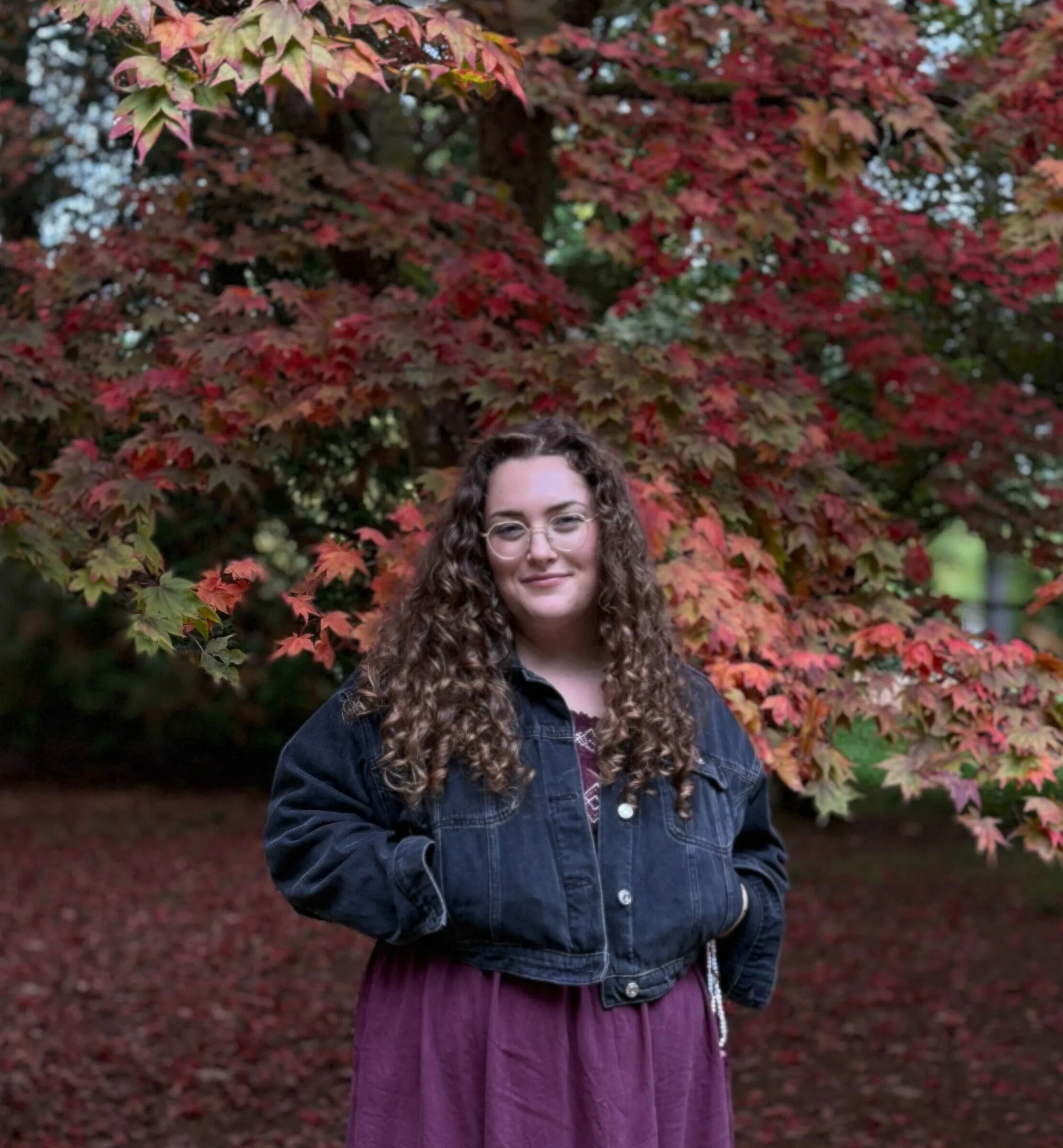 white woman in front of tree in the autumn, the leaves are turning red and pink and brown