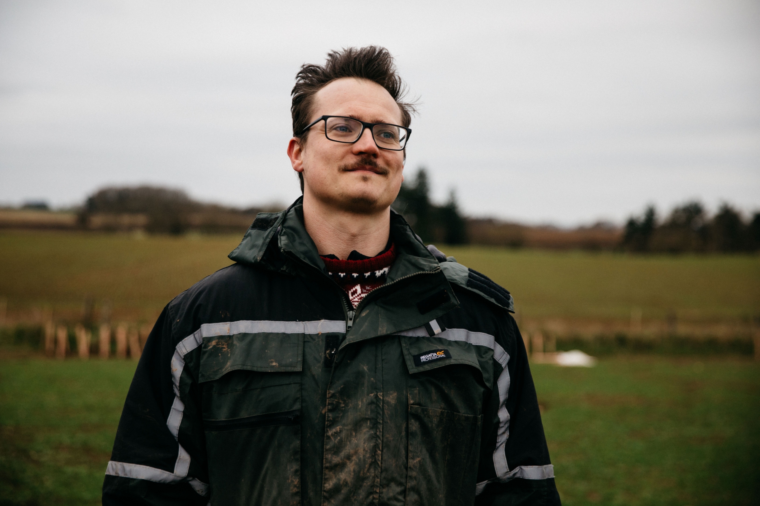 close up of white man in water proof jacket, behind is a field with trees